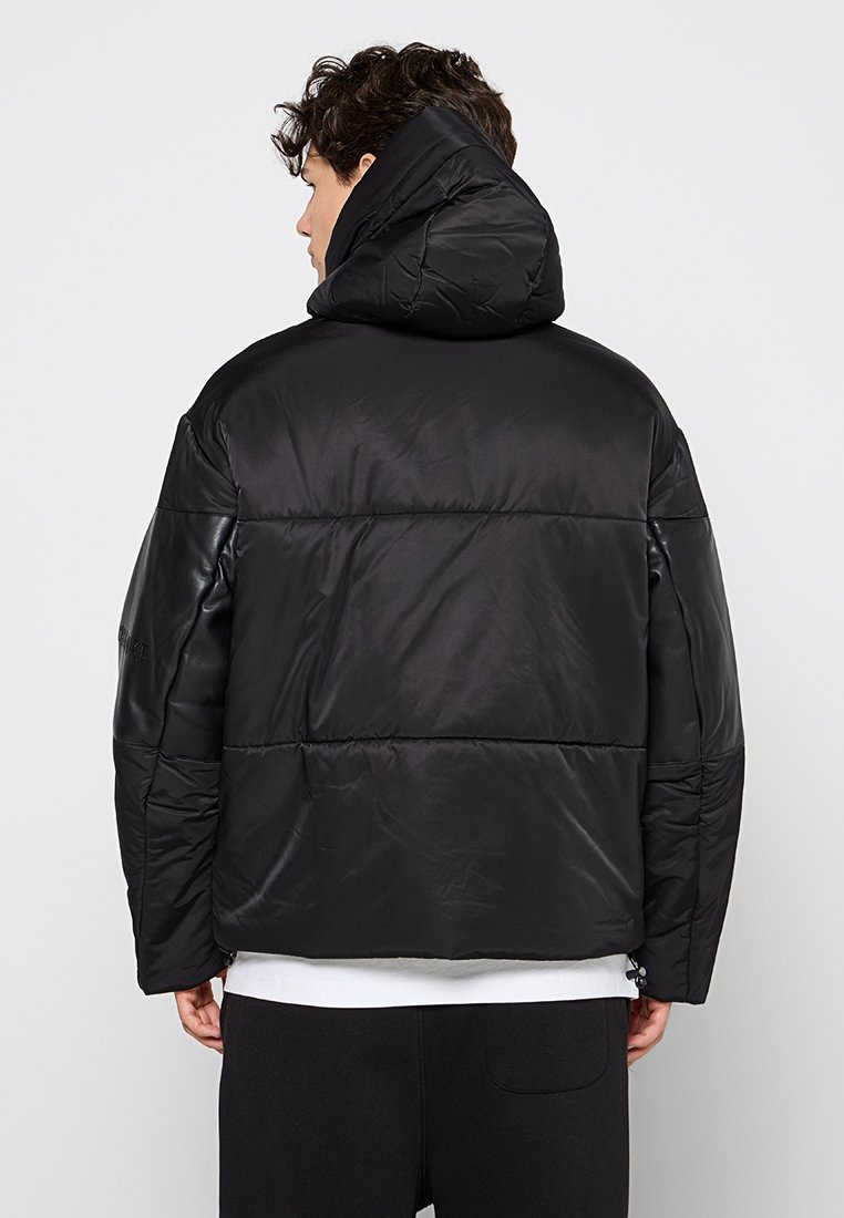 Giacca a vento nera con cappuccio, caratterizzata da una combinazione di texture opache e lucide, taglio dritto e dettagli di quilting visibili.