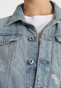 Jasnoniebieska kurtka dżinsowa o klasycznym kroju, z metalowymi zatrzaskami w kolorze srebrnym, zniszczonymi detalami, widocznymi szwami i dwiema kieszeniami na piersi.