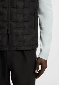 Svart quiltad väst med hög krage och dragkedjestängning, prydd med texturerat mönster och sidofickor med dragkedja. Buren över en ljusgrå långärmad skjorta.