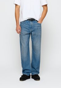 Zili džinsi ar brīvu piegriezumu, piecu kabatu dizainu un smalkiem nodilušiem akcentiem, apvienoti ar vienkāršu baltu T-kreklu un melnu jostu.