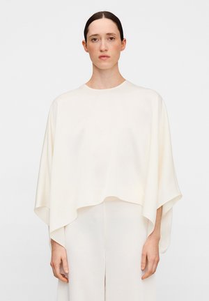 Vrouw met donker haar in het midden gescheiden, draagt een losse, gebroken witte poncho-achtige top met lange mouwen en bijpassende broek tegen een effen achtergrond.