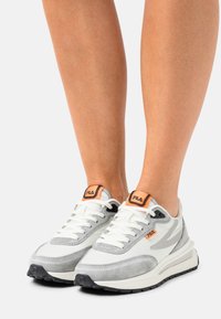 Jambes portant des baskets FILA blanches et grises avec des accents orange et des lacets blancs, debout sur un fond blanc.