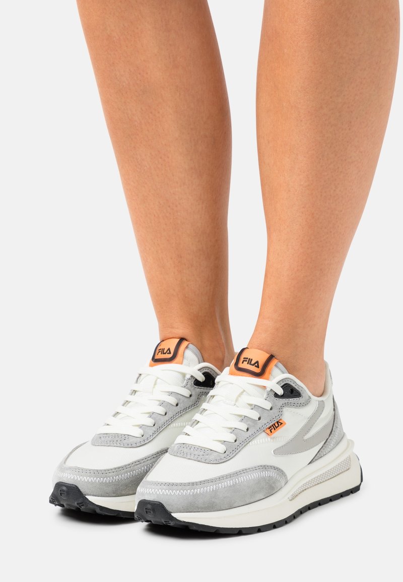 Jambes portant des baskets FILA blanches et grises avec des accents orange et des lacets blancs, debout sur un fond blanc.