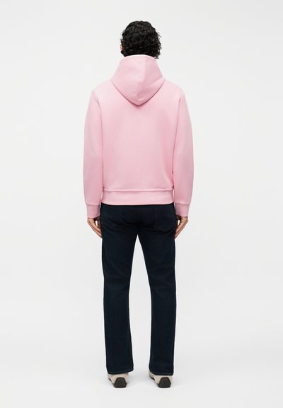 Άνδρας με σγουρά μαλλιά, που φοράει ροζ φούτερ με κουκούλα, σκούρο παντελόνι και ανοιχτά αθλητικά παπούτσια, στέκεται με την πλάτη προς το μέρος μας σε απλό λευκό φόντο.