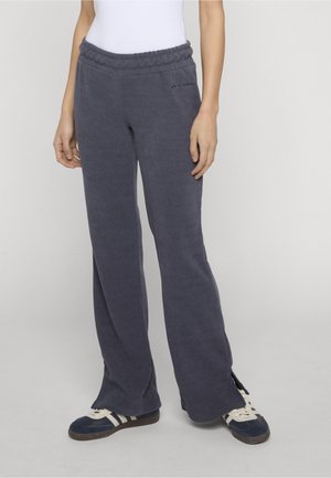 Persona vistiendo pantalones de pana gris oscuro holgados con cintura elástica y aberturas laterales, combinados con zapatillas deportivas azul marino y blancas.