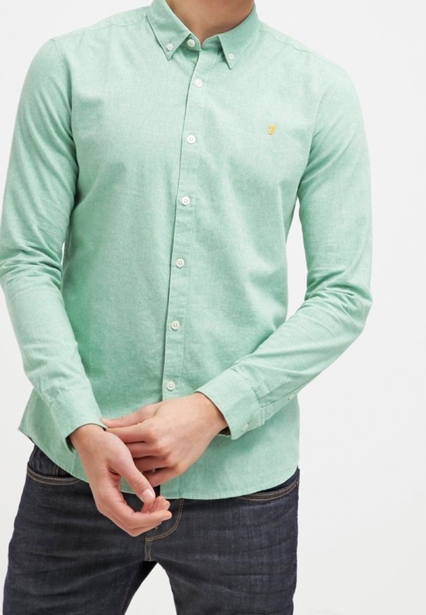 Homme portant une chemise à manches longues vert clair boutonnée avec un petit logo jaune sur la poitrine, associée à un jean bleu foncé, debout, les mains jointes.