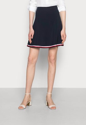 Kvinde iført en navyblå nederdel med hvid og rød stribet kant, hvid bluse med opkrogede ærmer og beige blokhøjhælede sandaler med ankelrem.