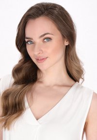 Portret osoby z długimi, falującymi brązowymi włosami, noszącej bezrękawnik w kształcie litery V w kolorze białym, na jednolitym białym tle.