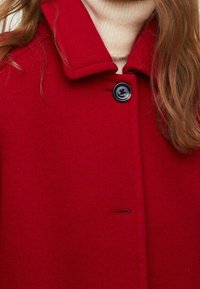 Femme portant un manteau rouge boutonné sur un pull à col roulé crème, avec des cheveux ondulés lâches visibles sur le côté droit.