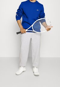 Modrý crewneck sweatshirt s logem, šedé tepláky a bílé tenisové boty, drží bílou tenisovou raketu s mřížkovaným vzorem.