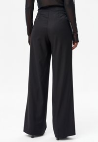 Osoba ubrana w czarne spodnie z wysokim stanem i szerokimi nogawkami oraz przezroczystą czarną bluzkę z długim rękawem, stojąca na białym tle.