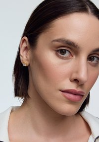 Złote kolczyki wkrętki z centralnym, przezroczystym kryształem, okrągły kształt, gładkie metaliczne wykończenie, noszone przez modelkę z krótkimi ciemnymi włosami.