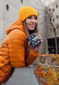 Černá peněženka s barevnými puntíky, kterou drží osoba v oranžové bundě a žluté pletené čepici, na pozadí venkovního prostředí.