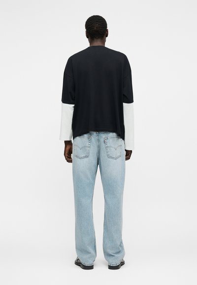 Άνδρας που φοράει μαύρο και άσπρο μακρυμάνικο πουκάμισο και ανοιχτόχρωμα μπλε χαλαρά τζιν, στέκεται με το πρόσωπο προς τα πίσω σε απλό φωτεινό φόντο.