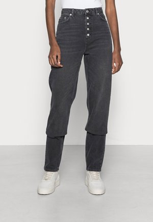 Person iført sorte højttaljede jeans med eksponeret knaplukning, lagdelt denimdesign over anklerne, hvide sneakers og en hvid top.