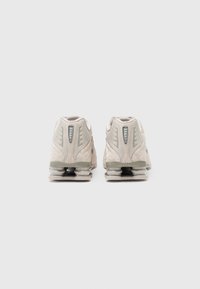 Para jasnobeżowych butów sportowych z widocznymi amortyzowanymi piętami, pokazanych z tyłu na jednolitym białym tle.