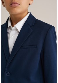 Marinblå kavaj med slät textur, enkelknäppt och med en bröstficka. Under syns en vit skjorta med krage.