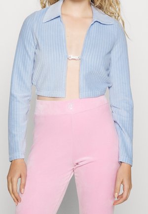 Femme portant une veste courte côtelée bleu clair avec un fermoir argenté sur une poitrine nue, associée à un pantalon en velours rose taille haute avec détail logo.