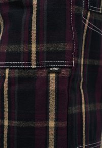 Κοντινό πλάνο υφάσματος καρό σε μπορντό, μαύρο και μπεζ με λευκή ραφή και μια μικρή μαύρη ετικέτα σε πτερύγιο τσέπης.