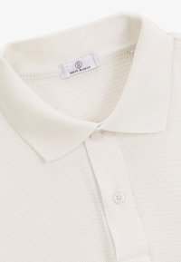 Polo bianco caratterizzato da un tessuto a maglia texturizzato, colletto classico e chiusura con due bottoni. Etichetta visibile all'interno del colletto.