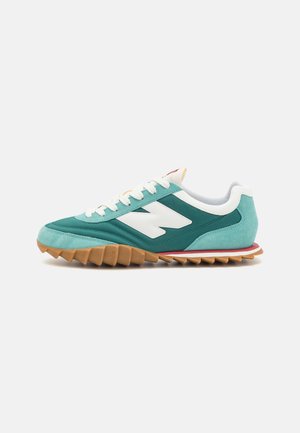 Chaussure de sport bleu sarcelle et turquoise avec lacets blancs, logo latéral blanc, et semelle en caoutchouc brune dentelée sur fond blanc.