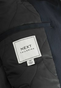 Mustvalge riie, millel on tepitud tekstuur, ja valge silt, millel on kiri "NEXT TAILORING" ning suurus "M". Õmmeldud silt, millel on kirjas "MADE IN CHINA."