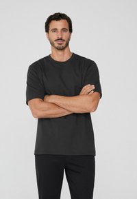 T-shirt noir à manches courtes, coupe décontractée, en tissu de coton mélangé doux. Le mannequin se tient les bras croisés devant un fond neutre.