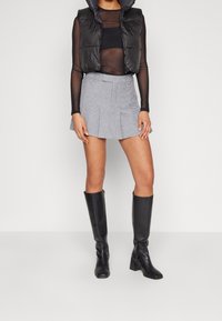 Veste sans manches matelassée noire, haut transparent à manches longues, mini-jupe plissée grise, et bottes noires hauteur genou avec embout carré. Textures lisses et design ajusté.