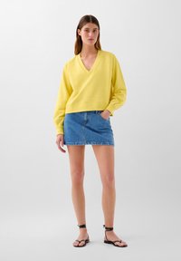 Pullover giallo con scollatura a V e orlo a costine, abbinato a una mini gonna in denim blu e sandali neri flat.