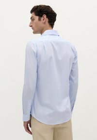 Camisa de botón de manga larga en azul claro con un cuello clásico, yugo trasero y puños abotonados. Presenta una tela suave y ligeramente texturada.