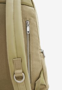 Béžový batoh s texturovaným tkaným tělem. Má stříbrný zip, boční kapsu a poutko na popruhu.