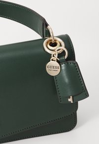 Green faux leather handbag with a structured shape. Features gold-tone hardware, a tag with "GUESS EST.1981," and contrasting stitching details.
