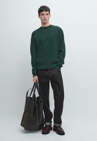 Dunkelgrüner, gerippter Pullover, graue Denim-Hose, braune Lederschuhe, mit einer dunklen Leder-Tasche. Das Modell steht vor einem neutralen Hintergrund.