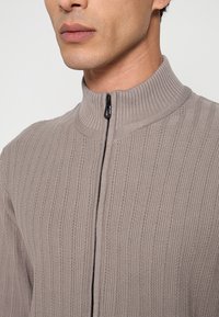 Taupekleurige gebreide trui met een hoge kraag, geribbelde textuur en een rits aan de voorkant. Heeft een slanke pasvorm en zachte stof.