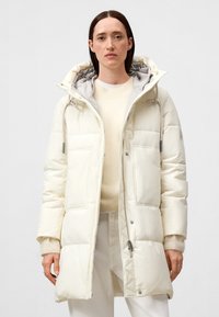 Kvinna i en krämfärgad puffjacka med huva, ljus tröja och vita byxor, stående framför en enfärgad bakgrund.