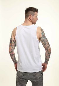 Camiseta sin mangas de algodón blanco con un corte relajado, escote redondo y detalle en el dobladillo trasero. Tatuajes visibles en los brazos en varios colores y diseños.