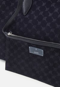 Granatowa torebka z teksturowanym zamszowym korpusem, wyposażona w zamek błyskawiczny, skórzane akcenty oraz widoczną metalową plakietkę z logo marki.