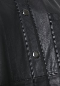 Vêtement en cuir noir avec deux boutons-pression métalliques visibles et une poche rectangulaire cousue sur le côté droit.