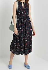 Abito floreale nero con un corpetto aderente, gonna a A e scollatura con fiocco; abbinato a scarpe blu chiaro e una borsa a spalla verde.