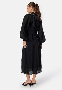 Robe noire semi-transparente avec des manches bouffantes et un col haut. Présente une taille smockée et un détail en nœud à l'arrière, descendant jusqu'au mollet.