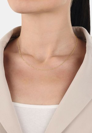 Collana in oro con un design delicato e attorcigliato. Indossata al collo, contrasta con l'abbigliamento chiaro e il tono della pelle.