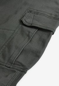 Dunkelolivgrüner Stoff, glatte Textur, mit einer Klappentasche und doppelter Naht entlang der Kanten. Fokus auf praktischen Design-Details.