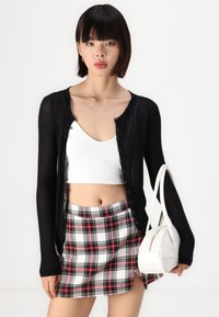 Črn dolgi kardigan z dolgimi rokavi in gumbi, bel crop top in karirana mini krila z rdečimi in črnimi poudarki. Mala bela torbica z zadrgo.