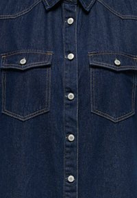 Tumši zila džinsa krekls ar sudrabotiem piespraužamiem pogām un divām krūšu kabatām. Raksturo kontrastējošas šuves un rāvējslēdzēja apkakles dizains.