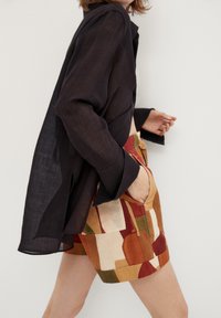 Czarny lniany koszula z długimi rękawami, zestawiona z wzorzystymi szortami, które prezentują abstrakcyjne kształty w stonowanych odcieniach pomarańczowego, zielonego i beżowego.