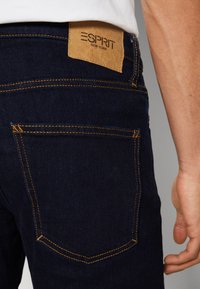 Vaqueros de mezclilla azul marino con costuras naranjas, mostrando una etiqueta de cuero en la cintura y un detalle en el bolsillo trasero.