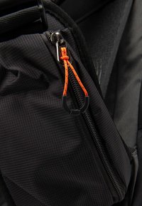 Coin d'un sac à dos en tissu noir avec une fermeture éclair munie d'un tirette orange et d'un accent en caoutchouc noir, mettant en valeur la texture du matériau et les détails du design.