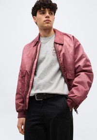 Νέος άνδρας που φοράει σατέν μπουφάν bomber σε χρώμα σκονιστό ρόζ πάνω από γκρι t-shirt με γραφικό σχέδιο και μαύρο παντελόνι με μαύρη ζώνη, στέκεται με το ένα χέρι στην τσέπη.