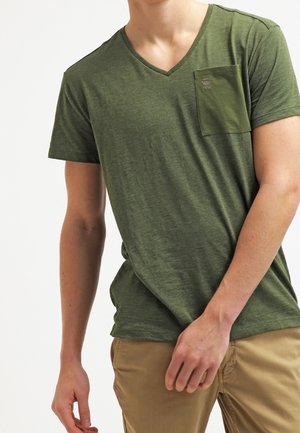 T-shirt col en V vert olive en tissu doux, avec une poche poitrine et un logo de marque discret. Texture douce et coupe décontractée.
