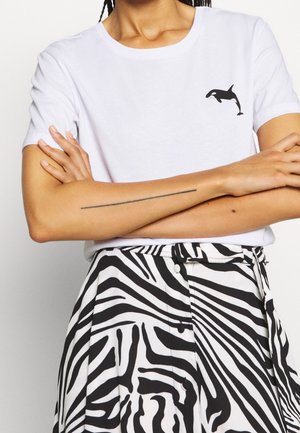 Persona con una camiseta blanca con diseño de orca negra y una falda con estampado de cebra en blanco y negro de cintura alta, brazos cruzados mostrando un tatuaje delgado en el brazo.